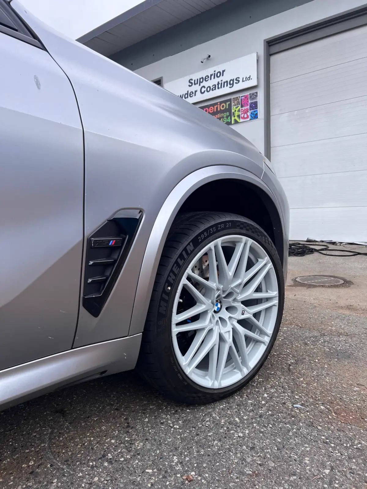 Silver BMW X5 M with frost white powder coated 21-inch wheels parked in front of the Superior Powder Coatings shop in North Vancouver